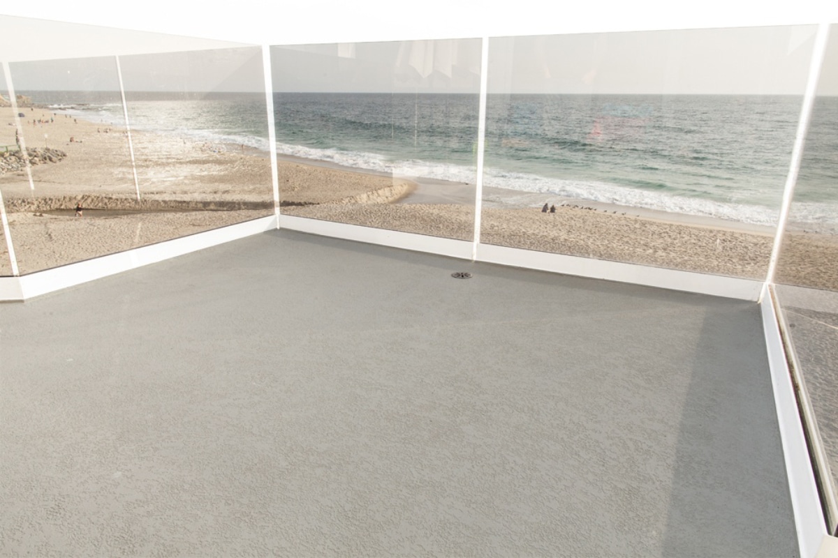 Rooftop patio with clear glass walls reflecting a sandy beach and ocean waves. Calm atmosphere with distant shoreline and soft overcast sky.